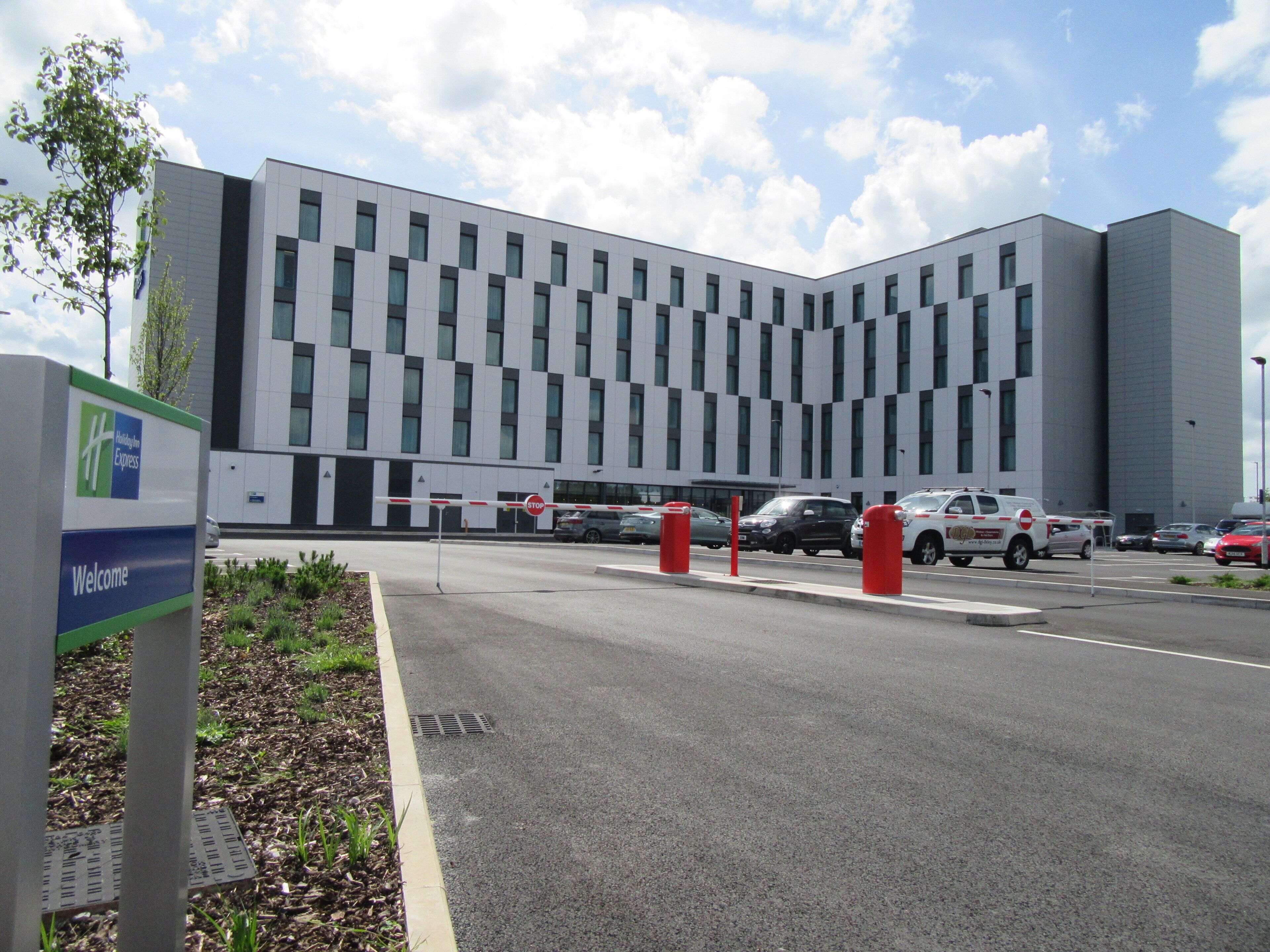 A modern building with a checkered facade, a 'Welcome' sign, and a gated parking area with vehicles.