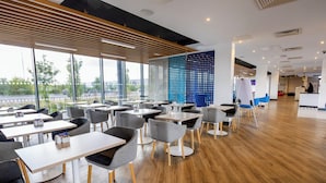 A modern restaurant with wooden flooring, grey and black chairs, and large windows offering a view of the street.