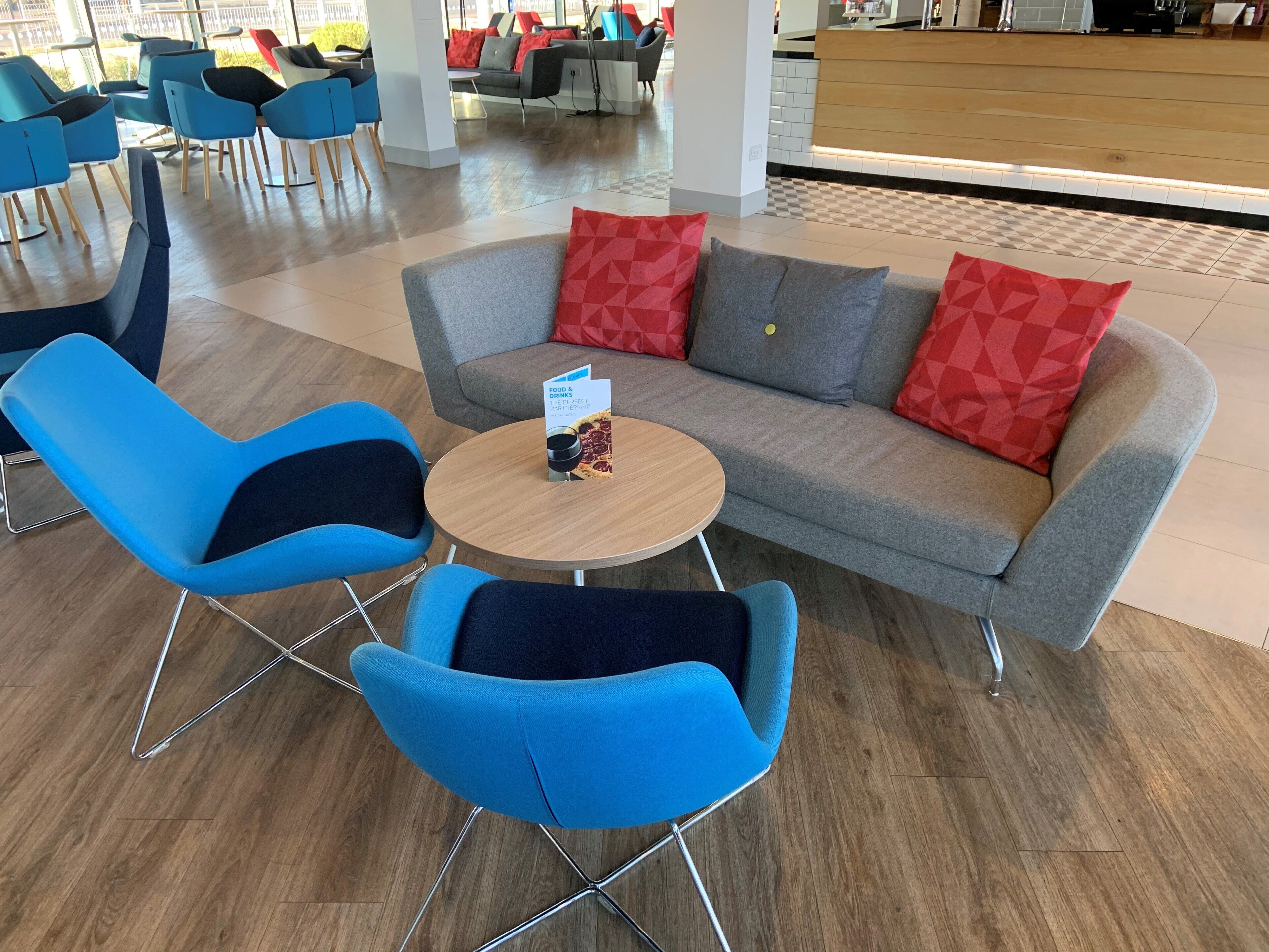 Ein stilvoller Loungebereich mit einem grauen Sofa, zwei blauen Sesseln und einem runden Holztisch mit einer Speisekarte.