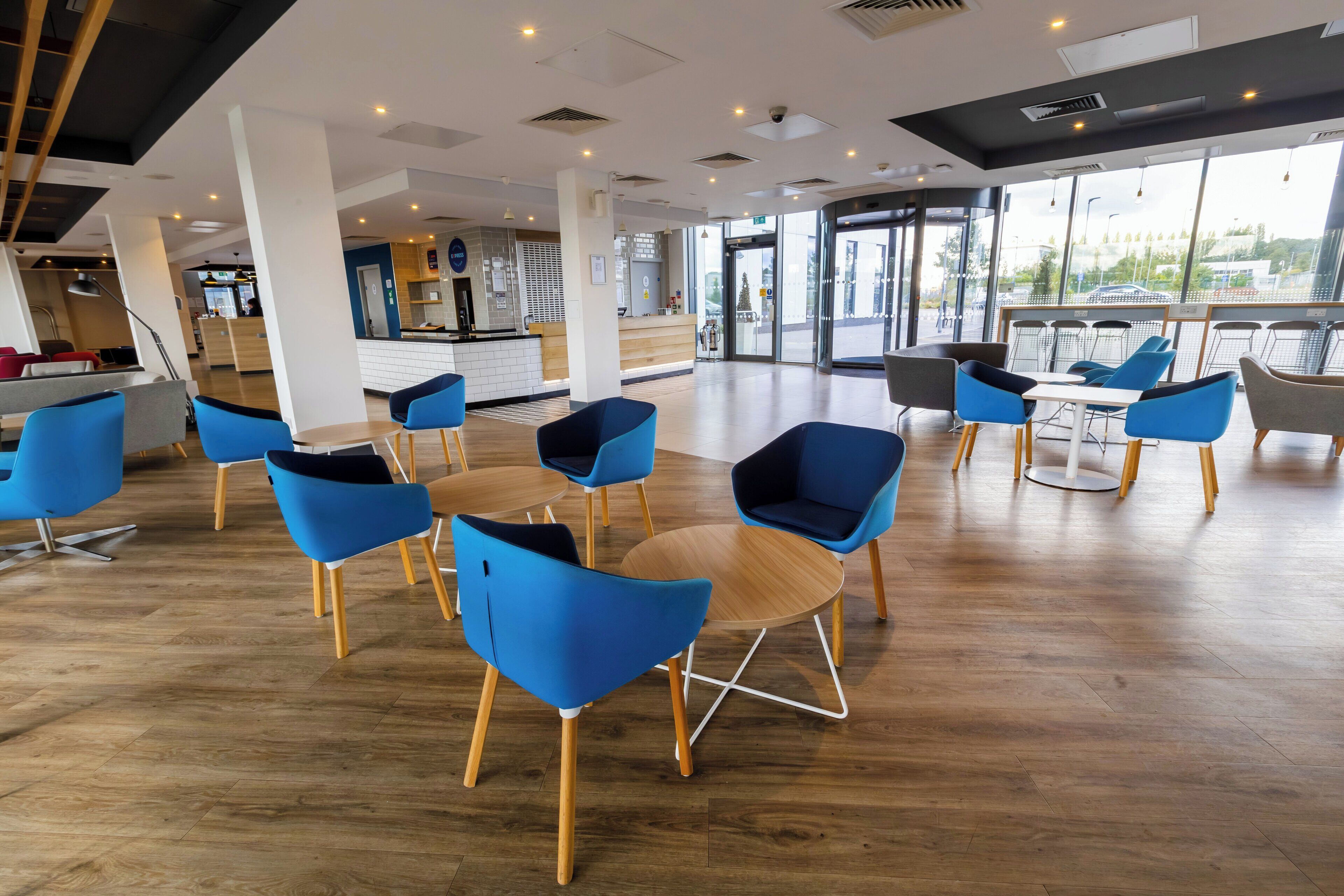 A modern interior with blue chairs and wooden tables, a reception area, and large windows offering a view outside.