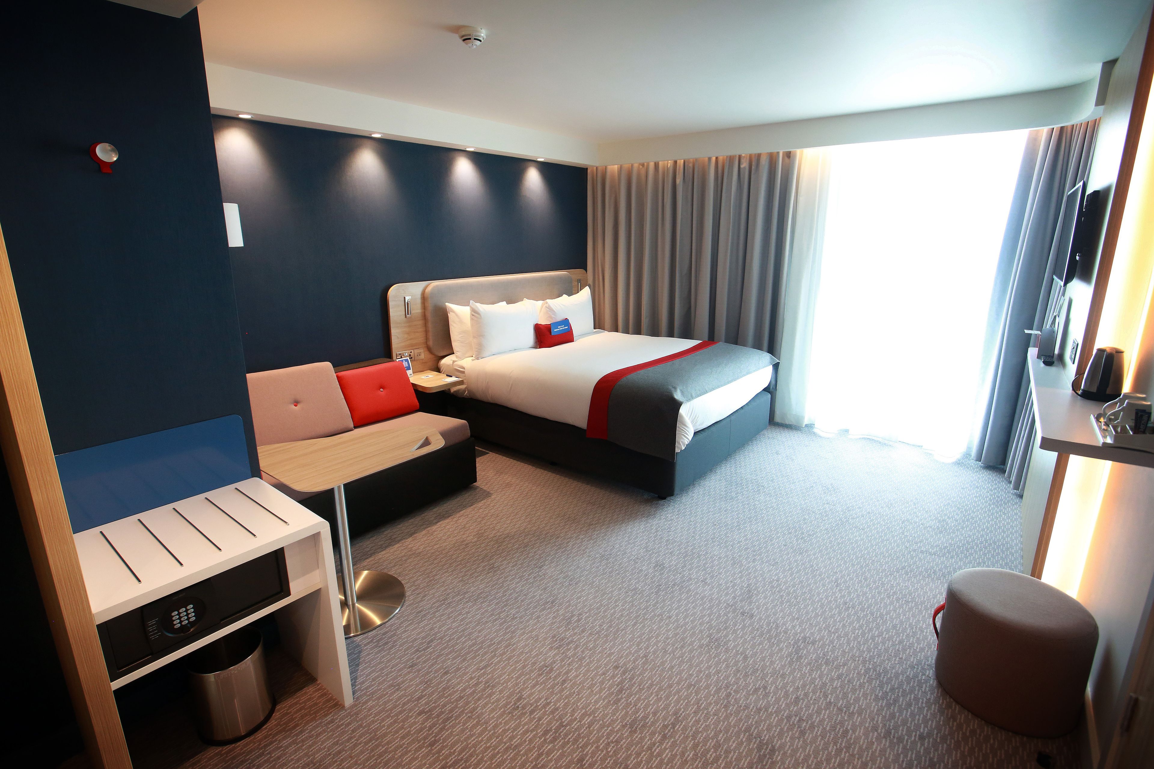 A hotel room with a bed, a desk, a chair, a trash bin, and a window with curtains.