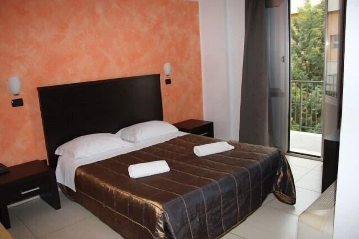 A hotel room with a large bed, two bedside tables, a nightstand, and a view of the outdoors through a sliding door.