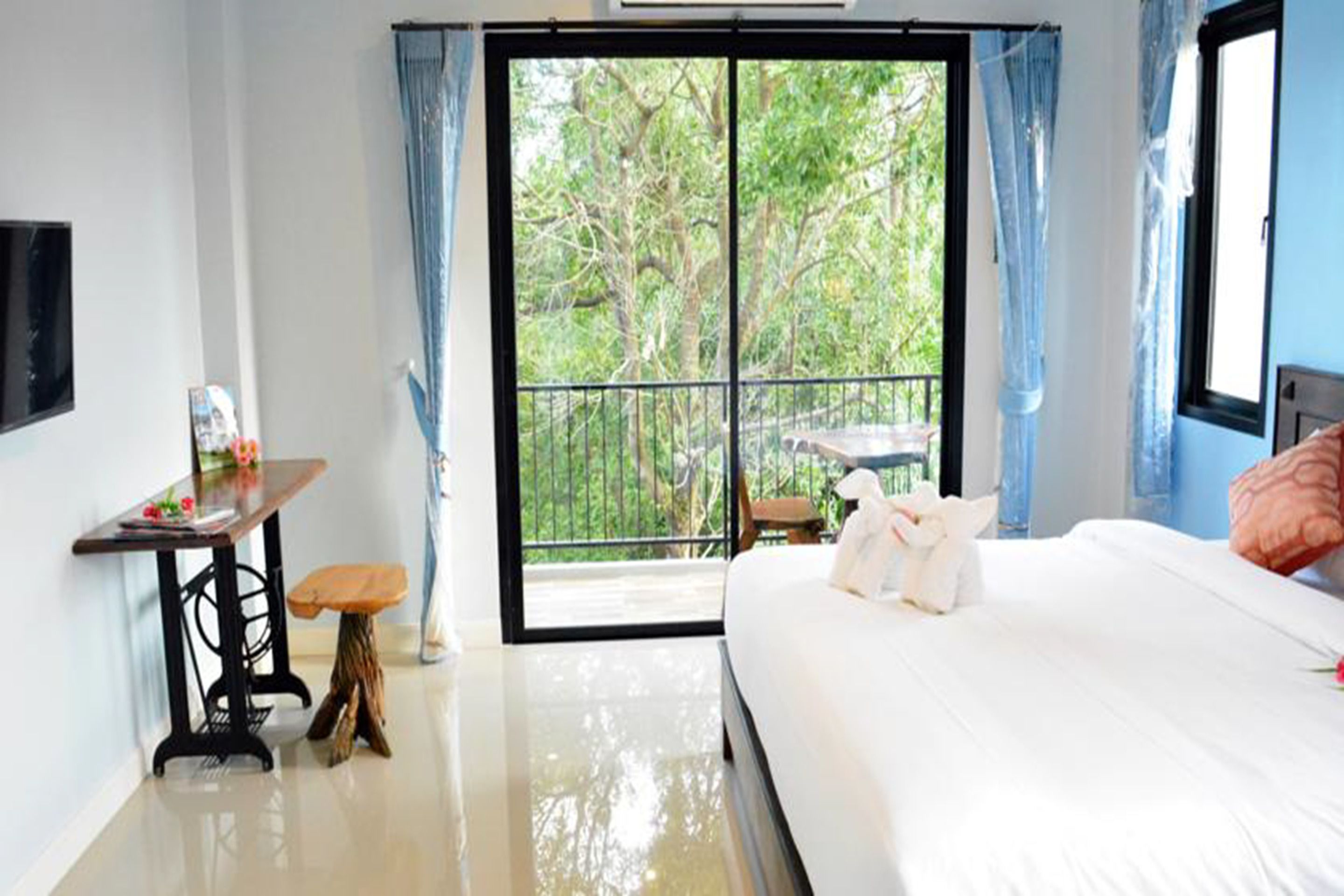 A hotel room with a bed, a small table, a bench, and a view of a balcony with trees.