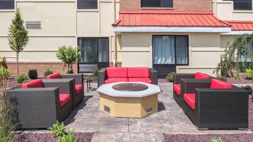 An outdoor seating area with wicker furniture and red cushions, a central fire pit, and a building with a red roof in the background.