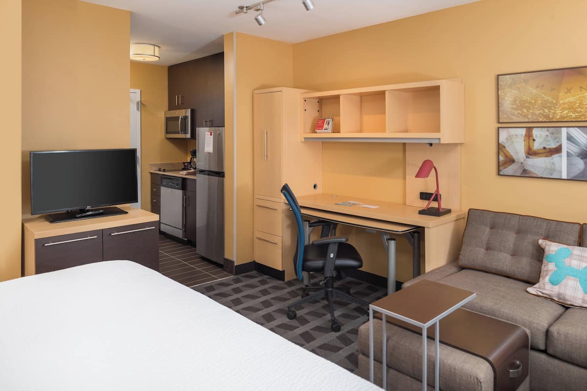 A hotel room with a bed, a desk with a chair, a kitchenette, a TV, and a sofa.