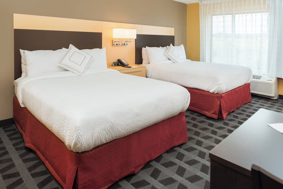 A hotel room with two beds, a nightstand with a phone, a lamp, and a window with curtains.