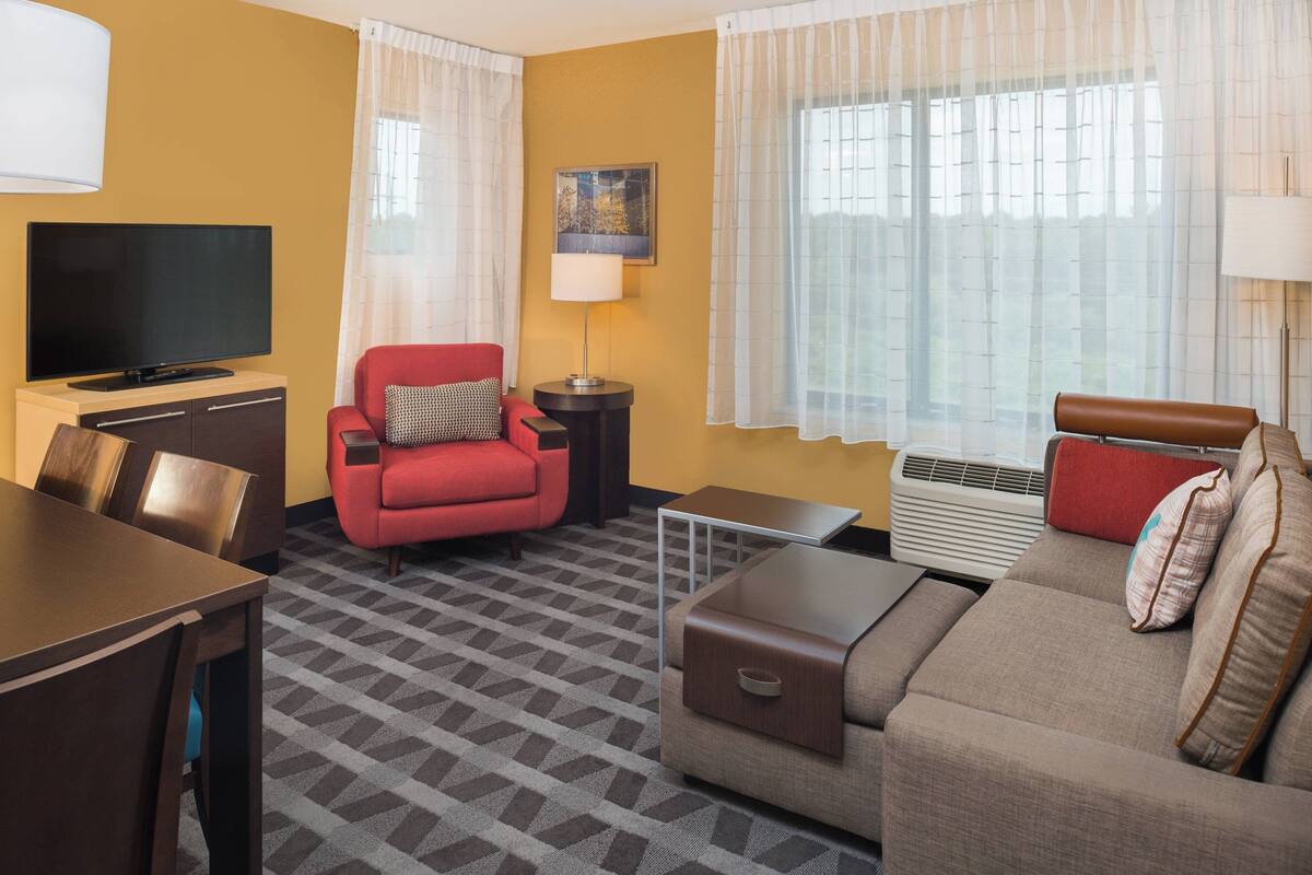 A hotel room with a TV, red armchair, sofa, dining table, and a patterned carpet.