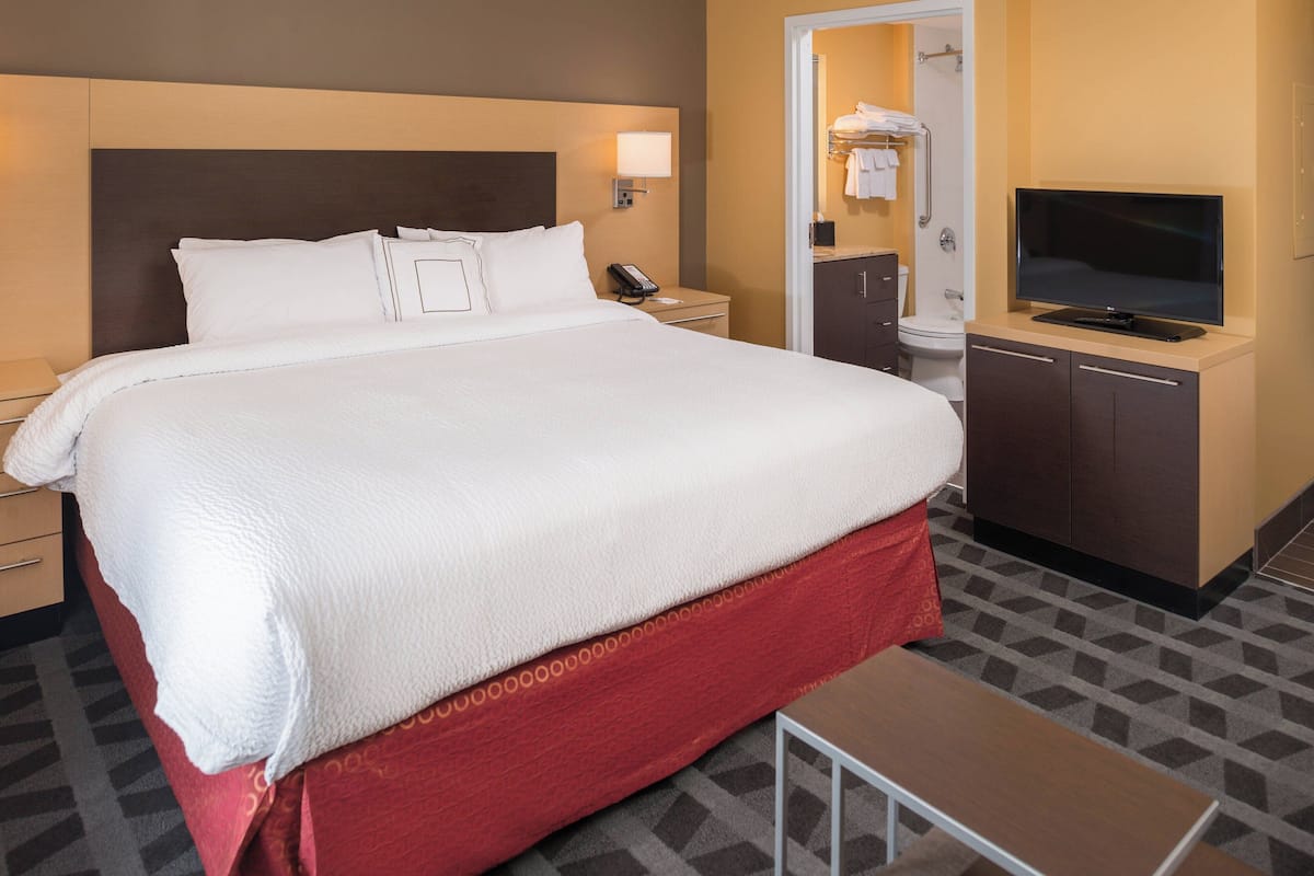 A hotel room with a large bed, bedside table, TV, and a bathroom visible through an open door.