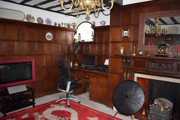 A traditional room with a wooden paneled wall, a fireplace, a desk with a computer, a television, and a red and white patterned rug. - Harmondsworth Hall Guest House Heathrow (West Drayton)