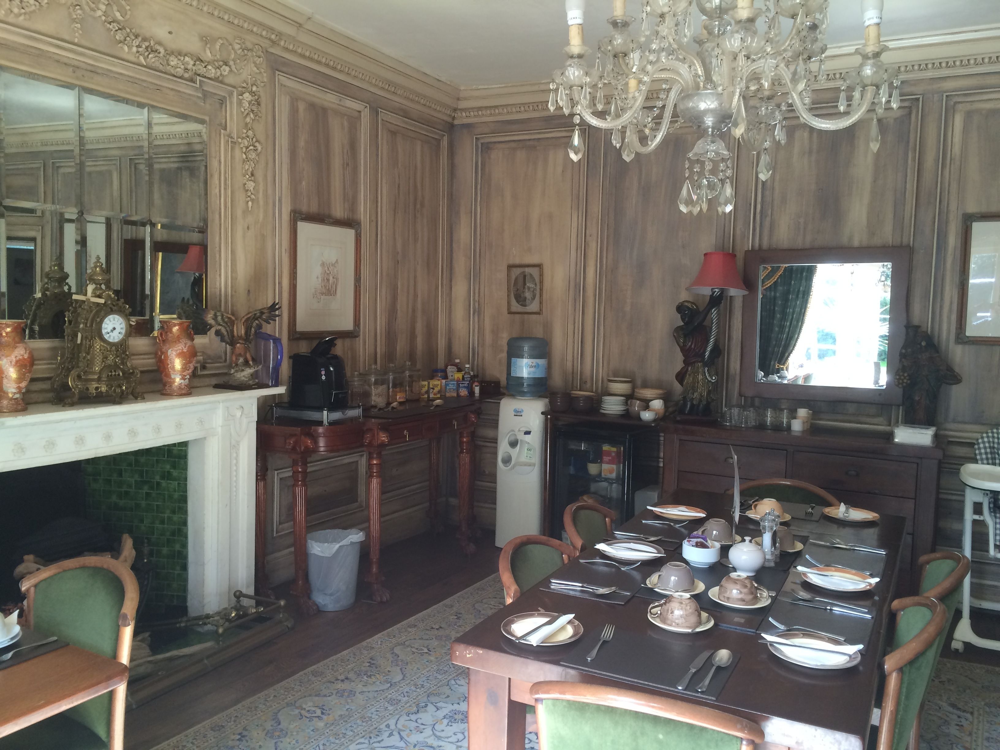 Uma sala de jantar com lustre, lambris de madeira, lareira e um espelho grande.