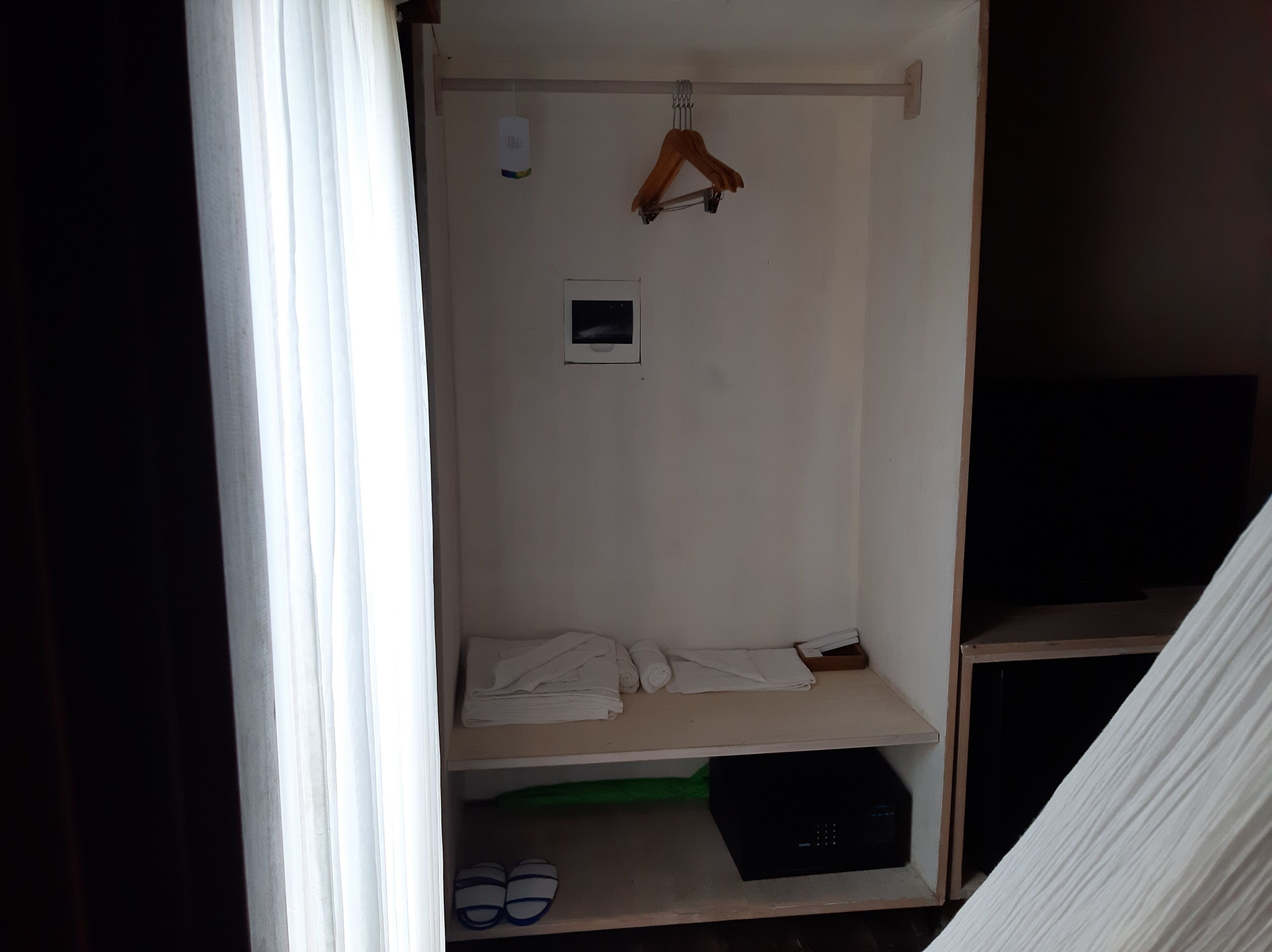 Une petite pièce faiblement éclairée, avec un lit simple, un porte-manteau suspendu et une petite étagère où sont pliés des vêtements et des chaussons.