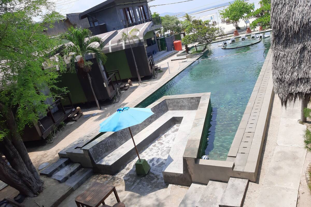 Une piscine accessible par des marches, entourée d’une terrasse en béton et surmontée d’une structure à toit de chaume.