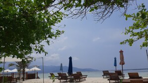 A beach with wooden lounge chairs and umbrellas, a sandy area, and a view of the sea and mountains in the background.