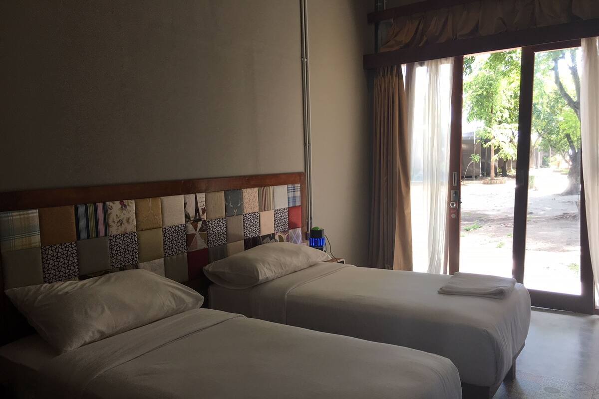 Une chambre d’hôtel avec deux lits, une tête de lit en bois ornée d’un motif patchwork, une table de chevet avec une lampe bleue et une vue sur l’extérieur grâce à de grandes fenêtres.