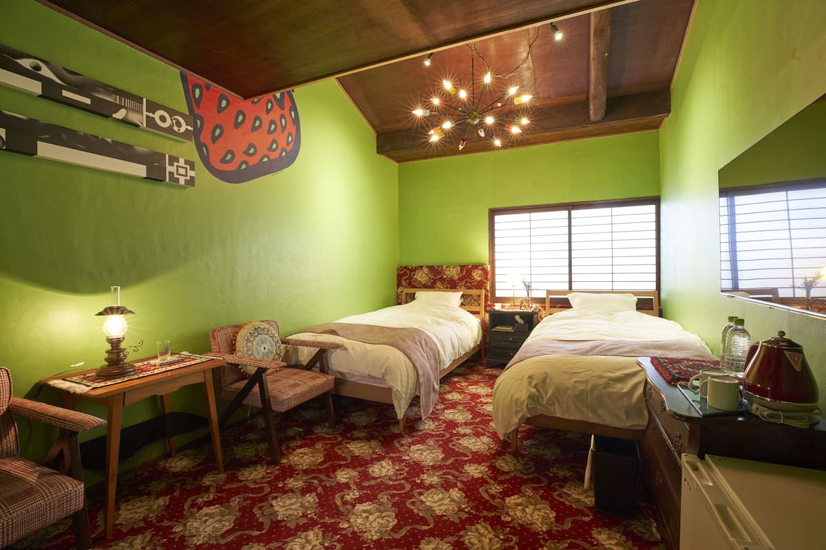 A room with two beds, a wooden table with a lamp, a chair, and a window with blinds.