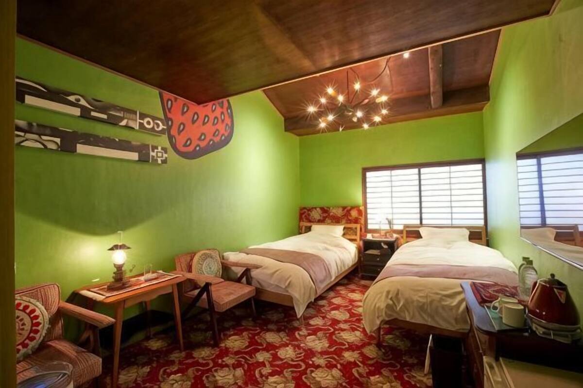 A room with two beds, a desk, a chair, a lamp, and a window with blinds.