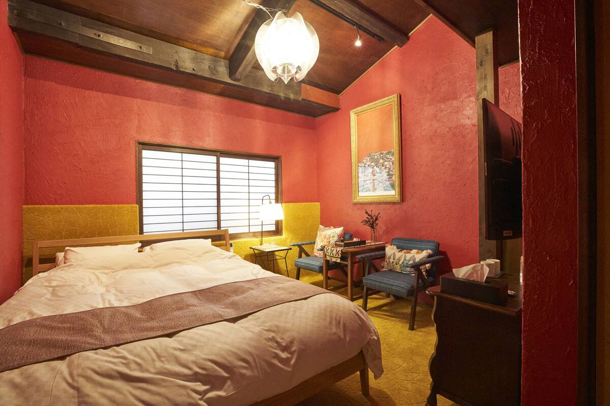 A room with a large bed, a desk with a chair, a window with blinds, and a painting on the wall.