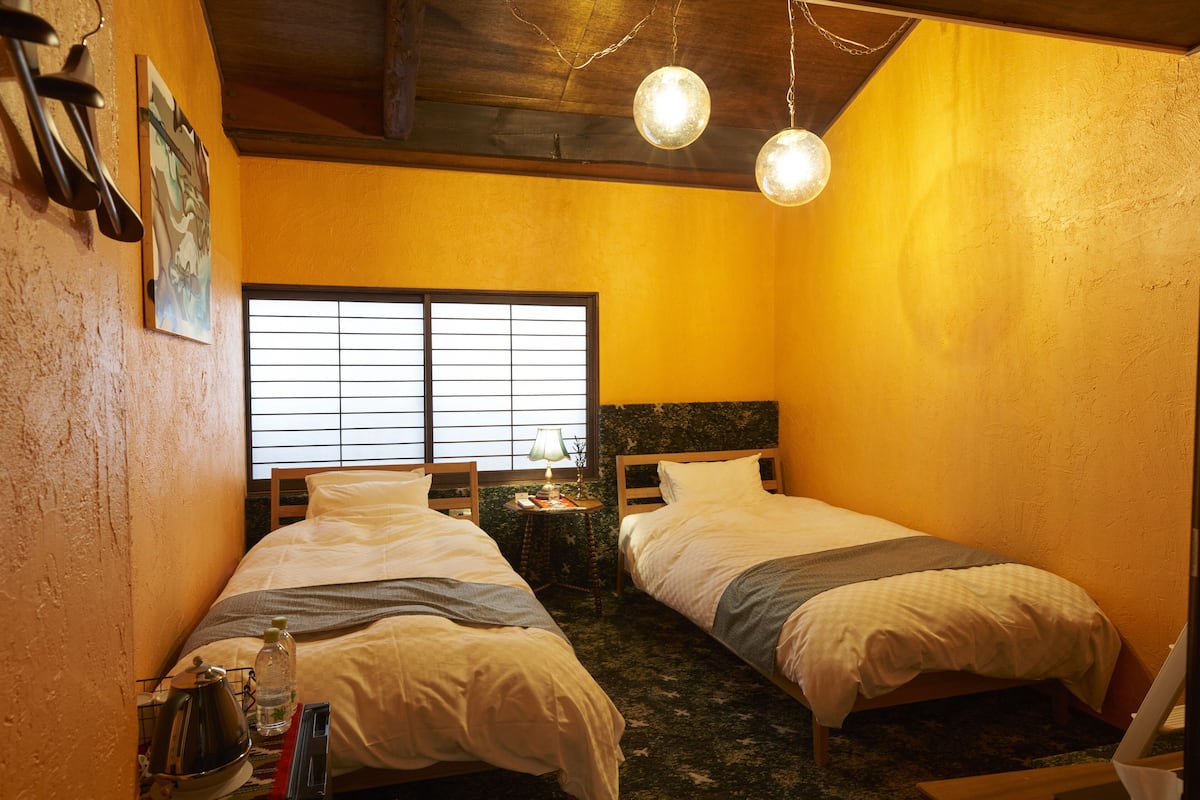 A small, cozy room with two beds, a window with blinds, a lamp on a side table, and a laptop on a desk.