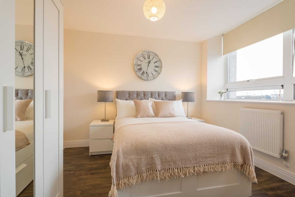 A bedroom with a bed, a nightstand, a wall clock, a window, and a radiator.