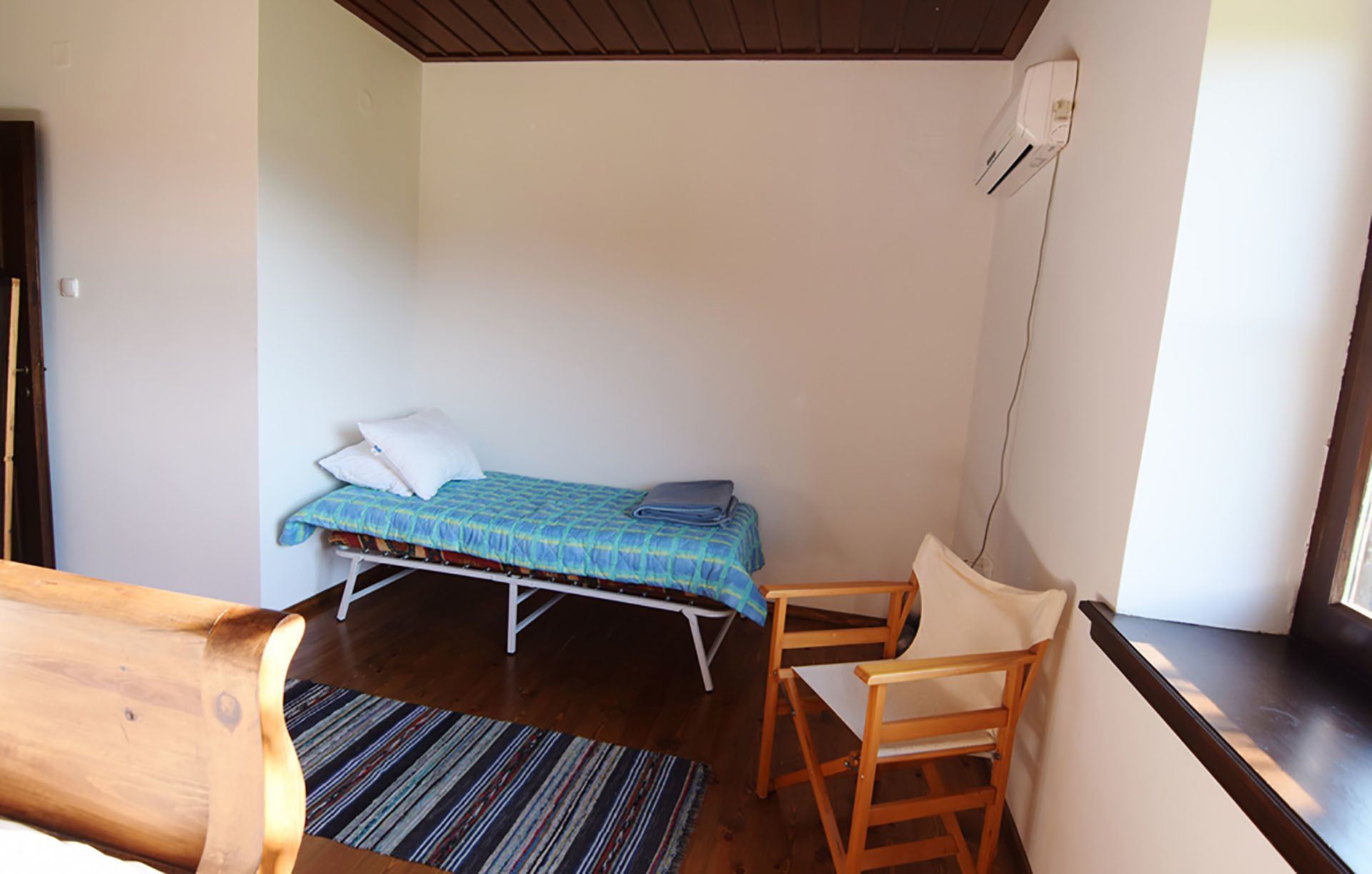 A small, single-bed room with a wooden desk and chair, a striped rug, and a white wall with a window.