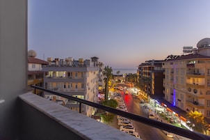 Una calle urbana al anochecer con edificios a ambos lados, una fila de coches aparcados y un restaurante con terraza visible.