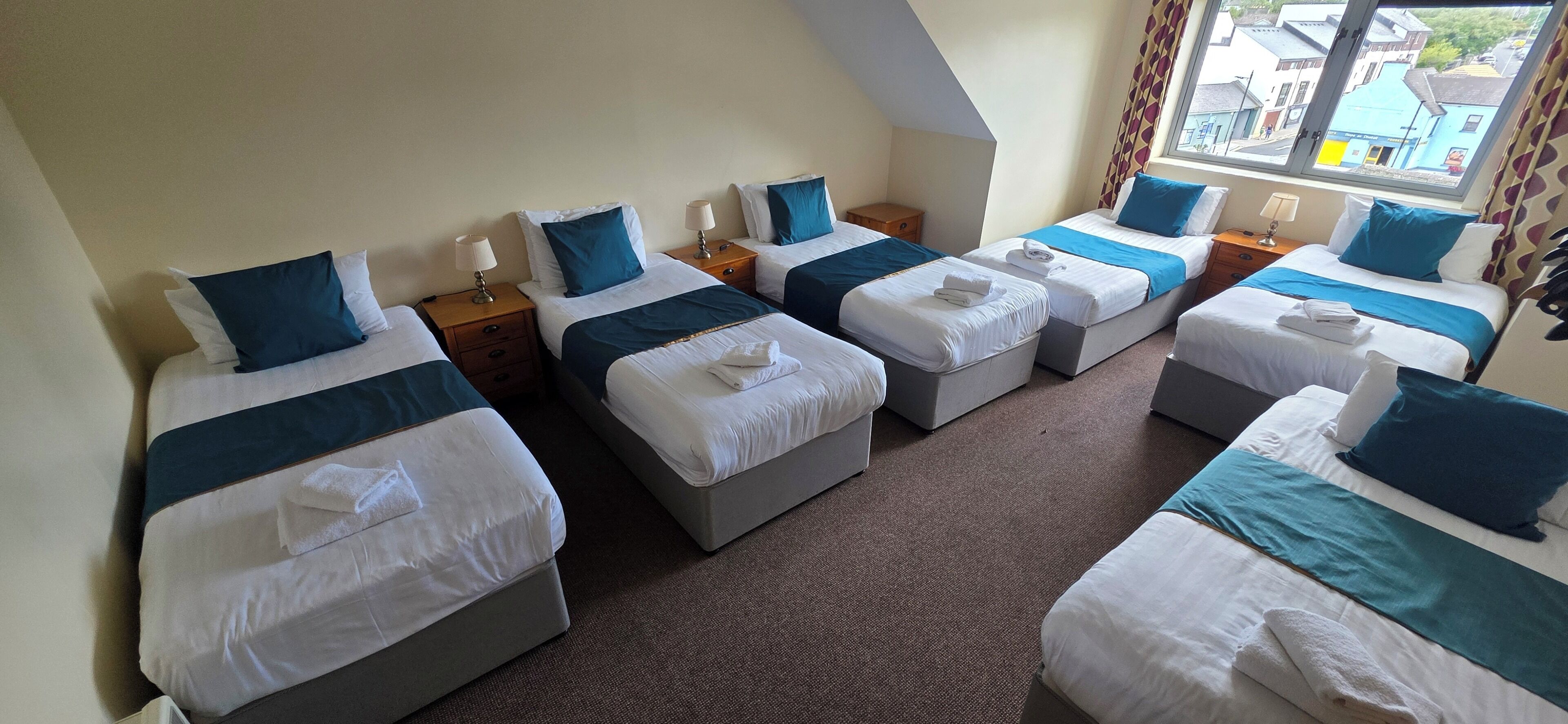 A hotel room with four single beds arranged in a row, each with a pillow, a bedside table, and a lamp. The room has a carpeted floor and a window with curtains.