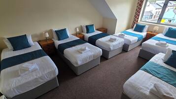A hotel room with four single beds arranged in a row, each with a pillow, a bedside table, and a lamp. The room has a carpeted floor and a window with curtains.