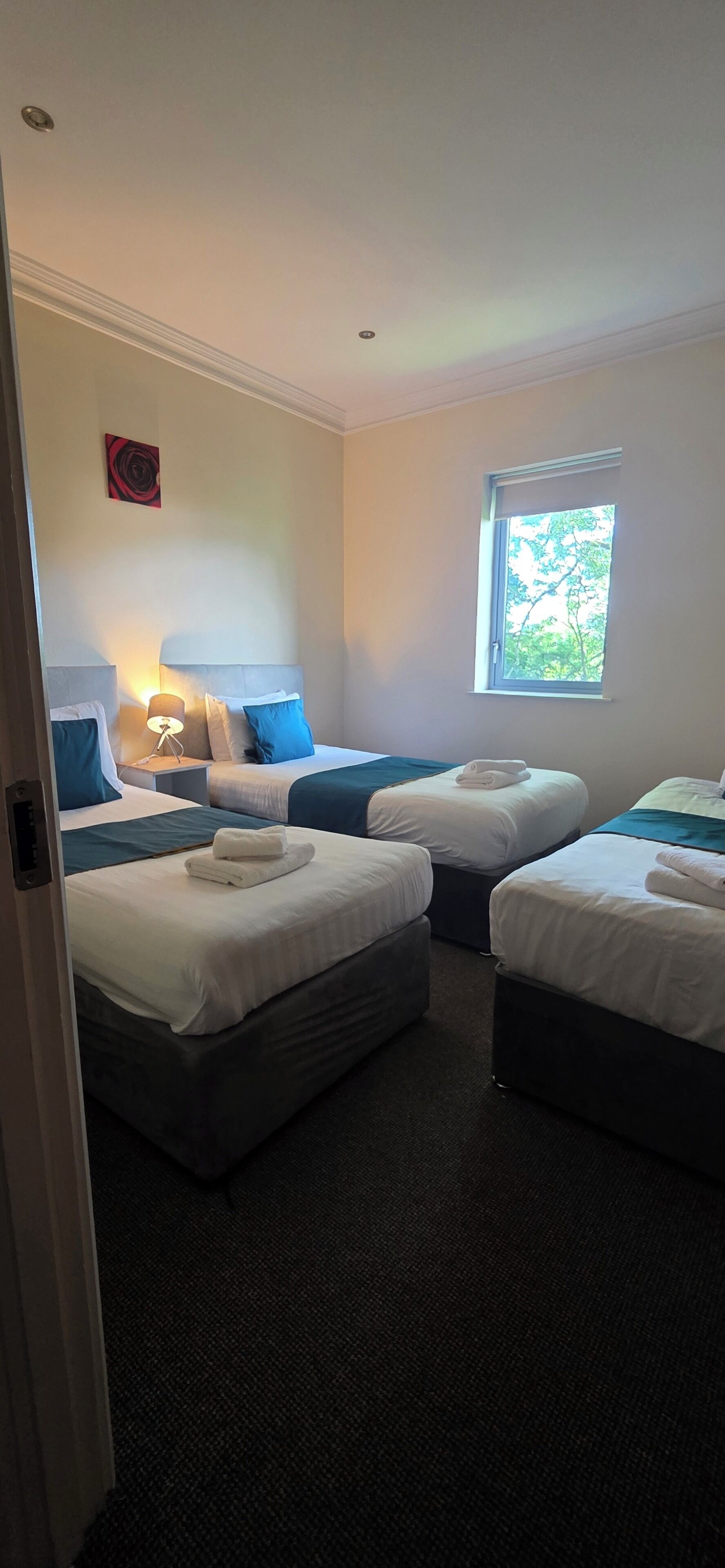 A hotel room with three beds, a window with a view of trees, a painting on the wall, and a lamp on the nightstand.