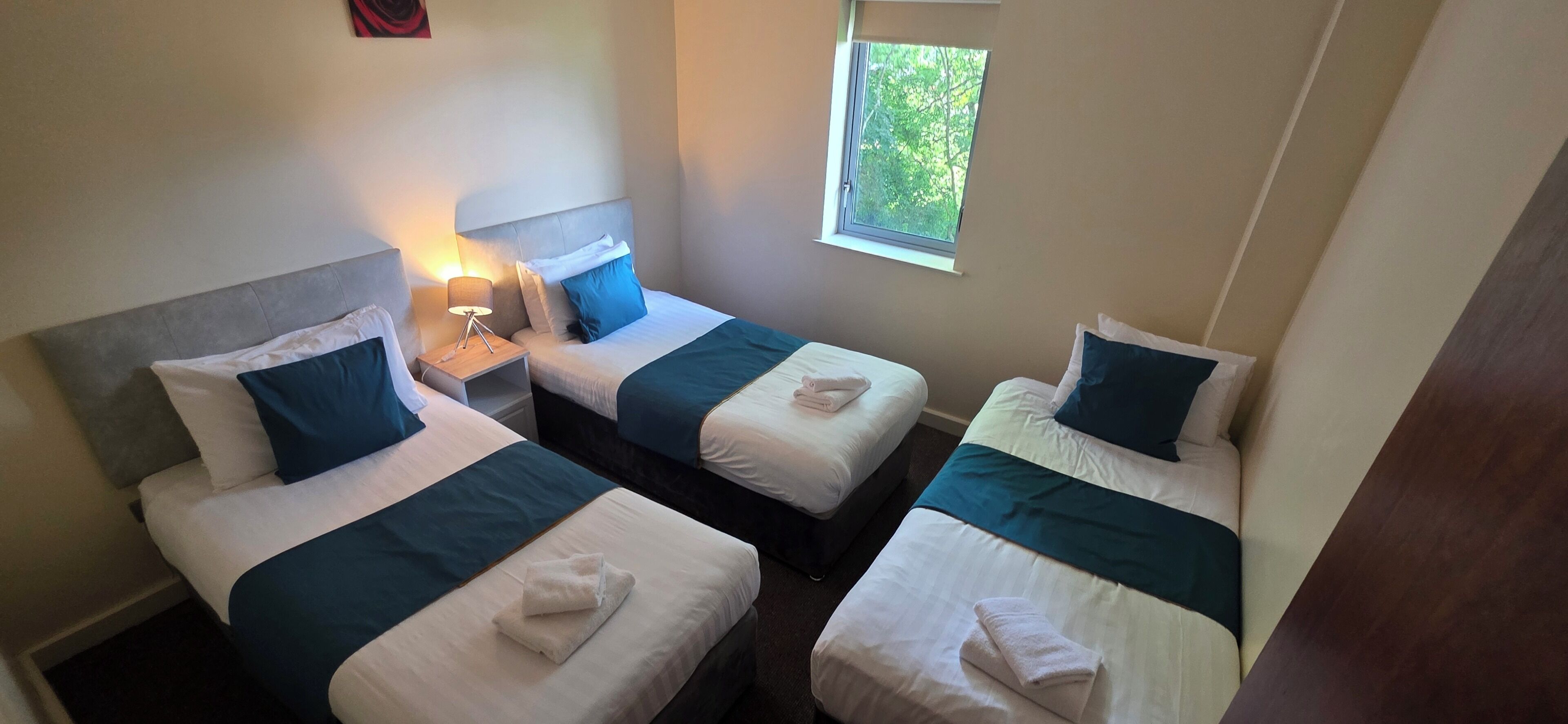 Une chambre d’hôtel avec trois lits simples, chacun agrémenté d’une touche de couleur bleue, une table de chevet avec une lampe et une fenêtre donnant sur un paysage verdoyant.
