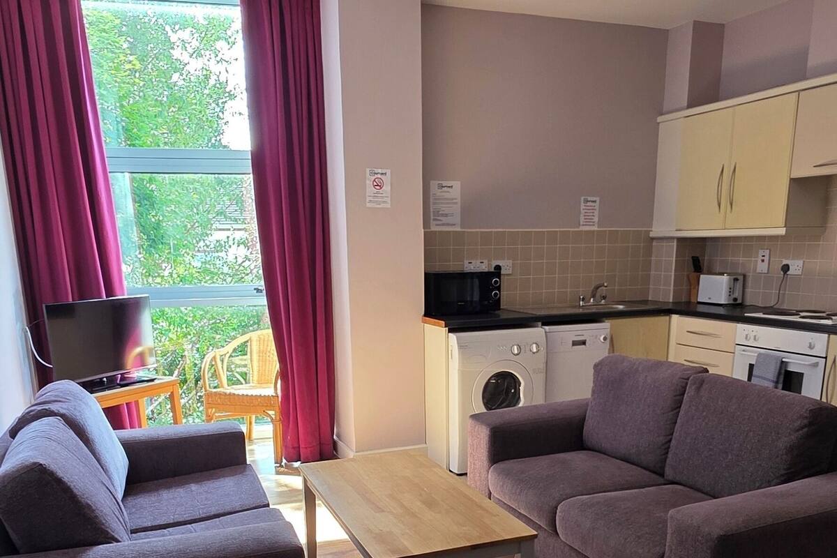 Ein modernes Wohnzimmer mit einer Couch, einem Couchtisch und einem Fernseher. Es gibt eine Küchenzeile mit Schränken und einer Mikrowelle.