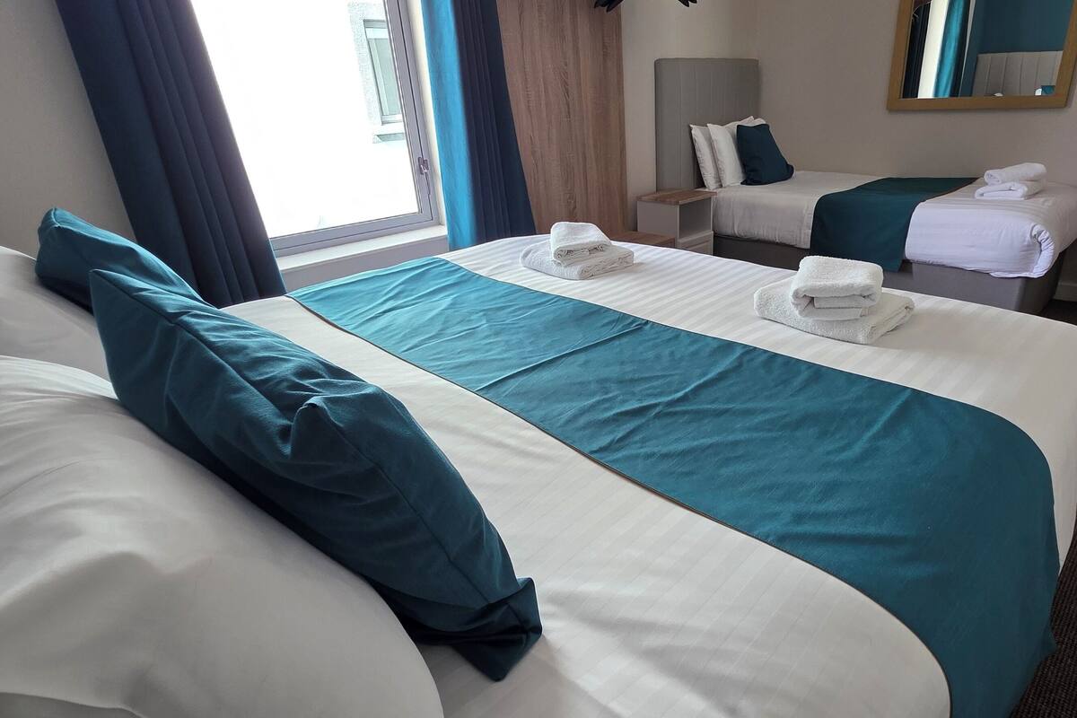 Ein Hotelzimmer mit einem großen Bett, einem kleineren Bett und einem Schreibtisch. Auf den Betten liegen Handtücher und an der Wand befindet sich ein Spiegel.