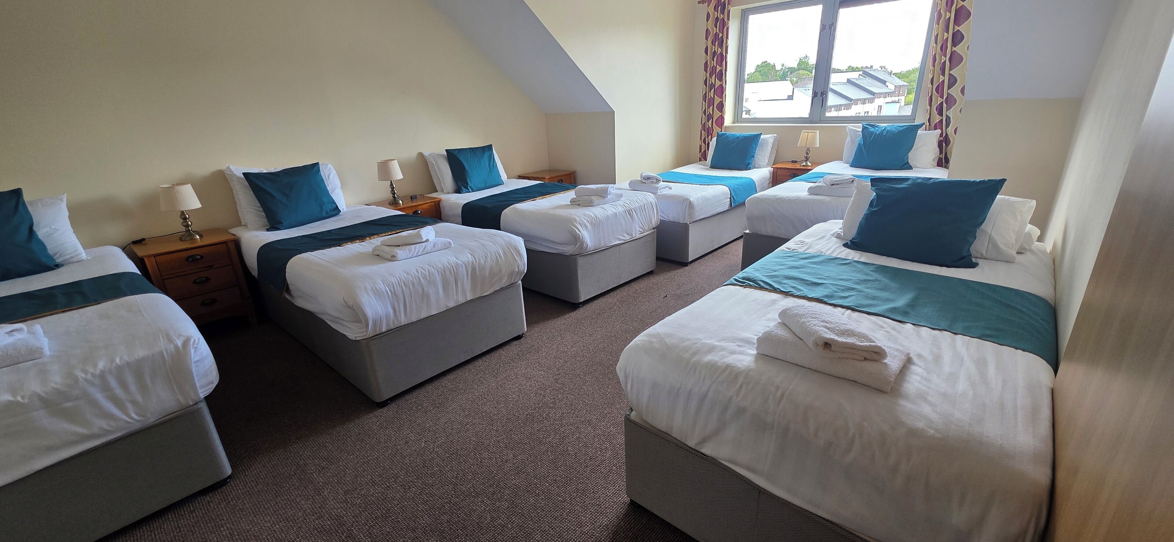 A hotel room with four beds arranged in a U-shape, each with white bedding and blue pillows. There are bedside tables with lamps and a window with curtains.