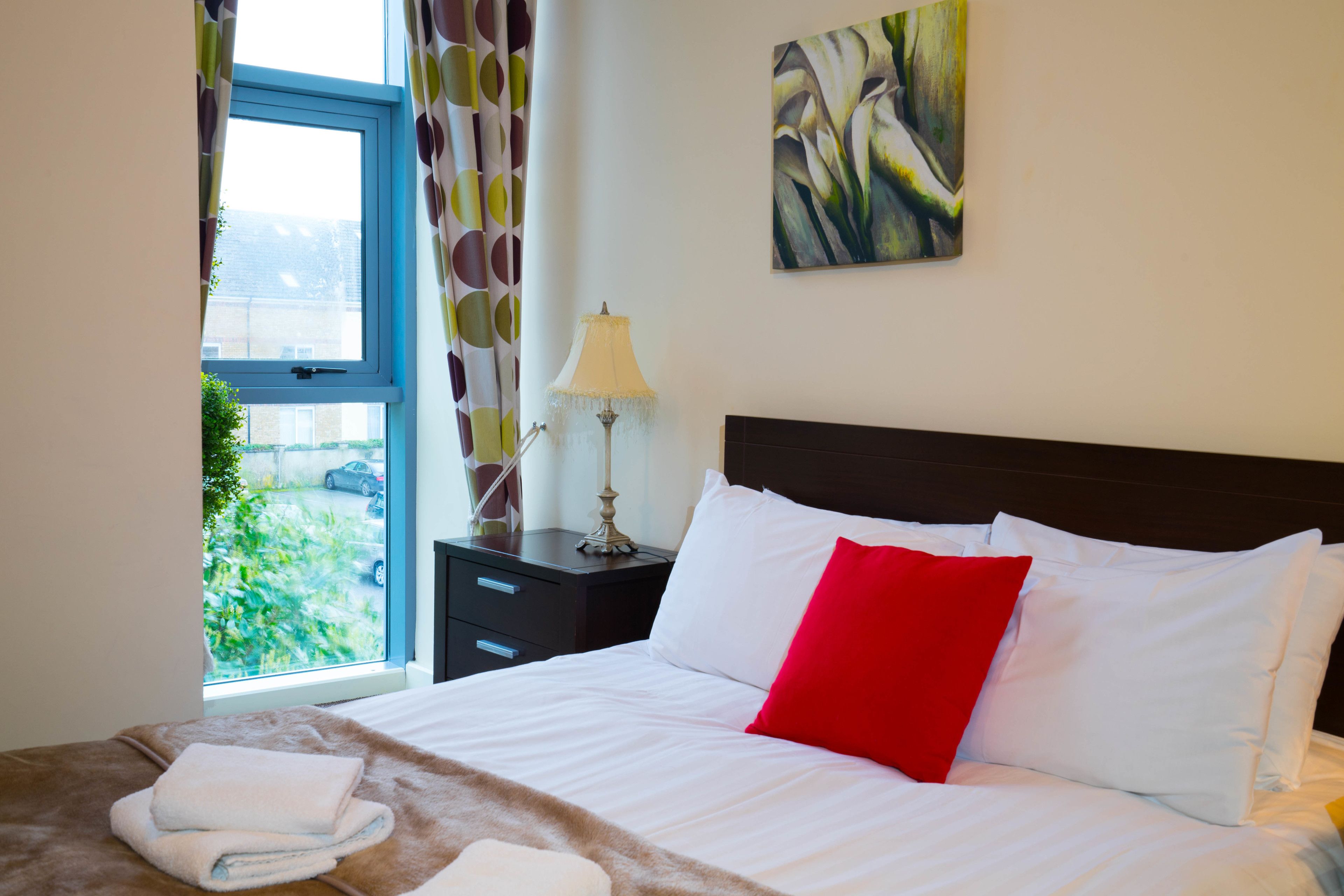 A neatly made bed with white and red pillows, a lamp on a bedside table, and a window with curtains.