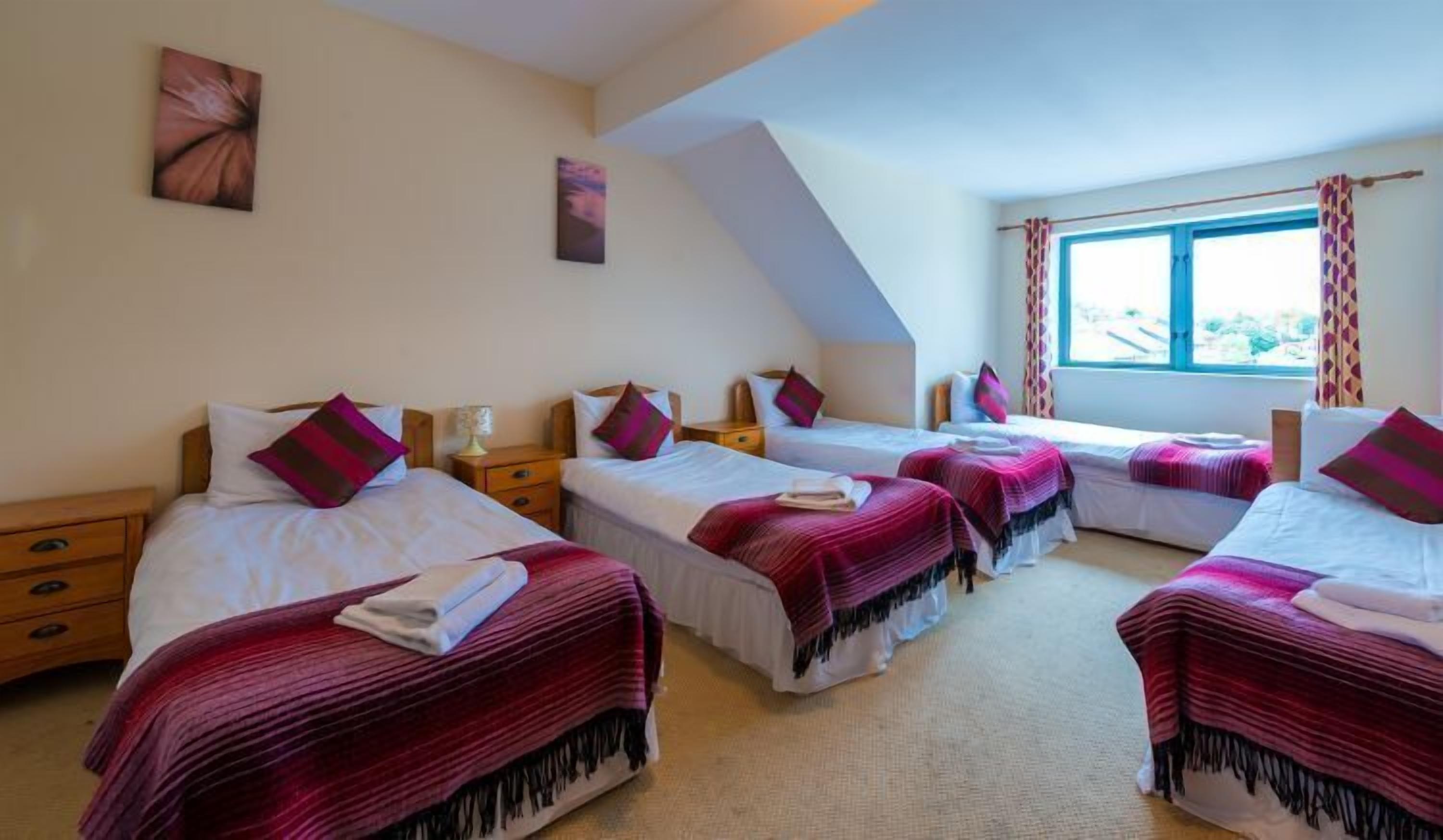 Habitación de hotel con cuatro camas, cada una con cubrecama y almohadas rojas y blancas. Hay mesitas de noche de madera con lámparas, un ventanal con cortinas y obras de arte en las paredes.