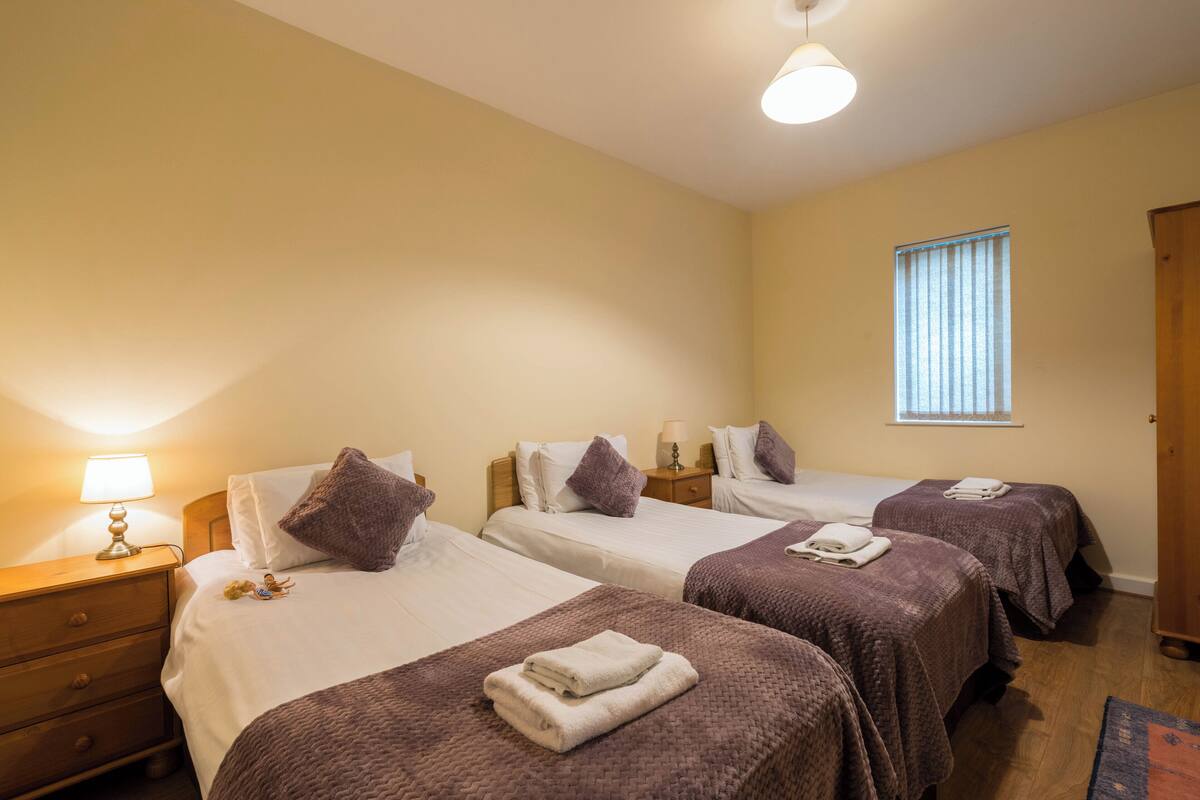 Ein Hotelzimmer mit drei Betten, jeweils mit brauner Bettdecke und weißen Laken. Es gibt ein Fenster mit Jalousien, einen Holzschrank und einen Nachttisch mit einer Lampe.