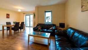A living room with a leather couch, a glass coffee table, a dining table with chairs, and a television on a stand. - The Courtyard Apartments (Carrick-on-Shannon)