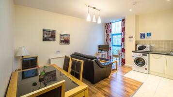 A compact living space with a sofa, a dining table with chairs, a TV, a washing machine, and a window with curtains.