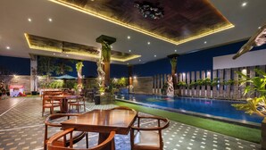 Zona de piscina con mesas y sillas de madera, una piscina y plantas decorativas.