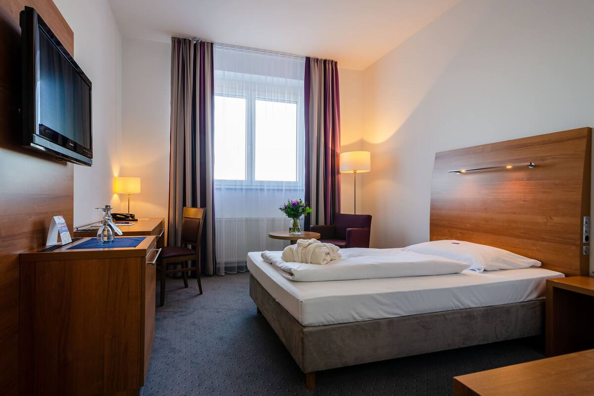 A hotel room with a bed, a desk, a chair, a TV, and a window with curtains.