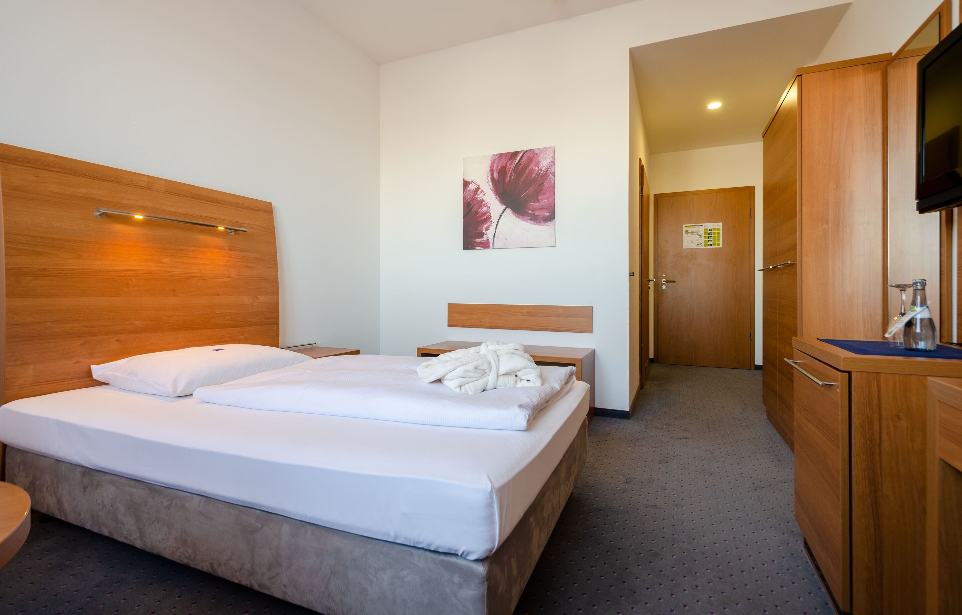 A hotel room with a wooden headboard, a bed with white linens, a wooden nightstand, a flat-screen TV mounted on the wall, and a wooden wardrobe.