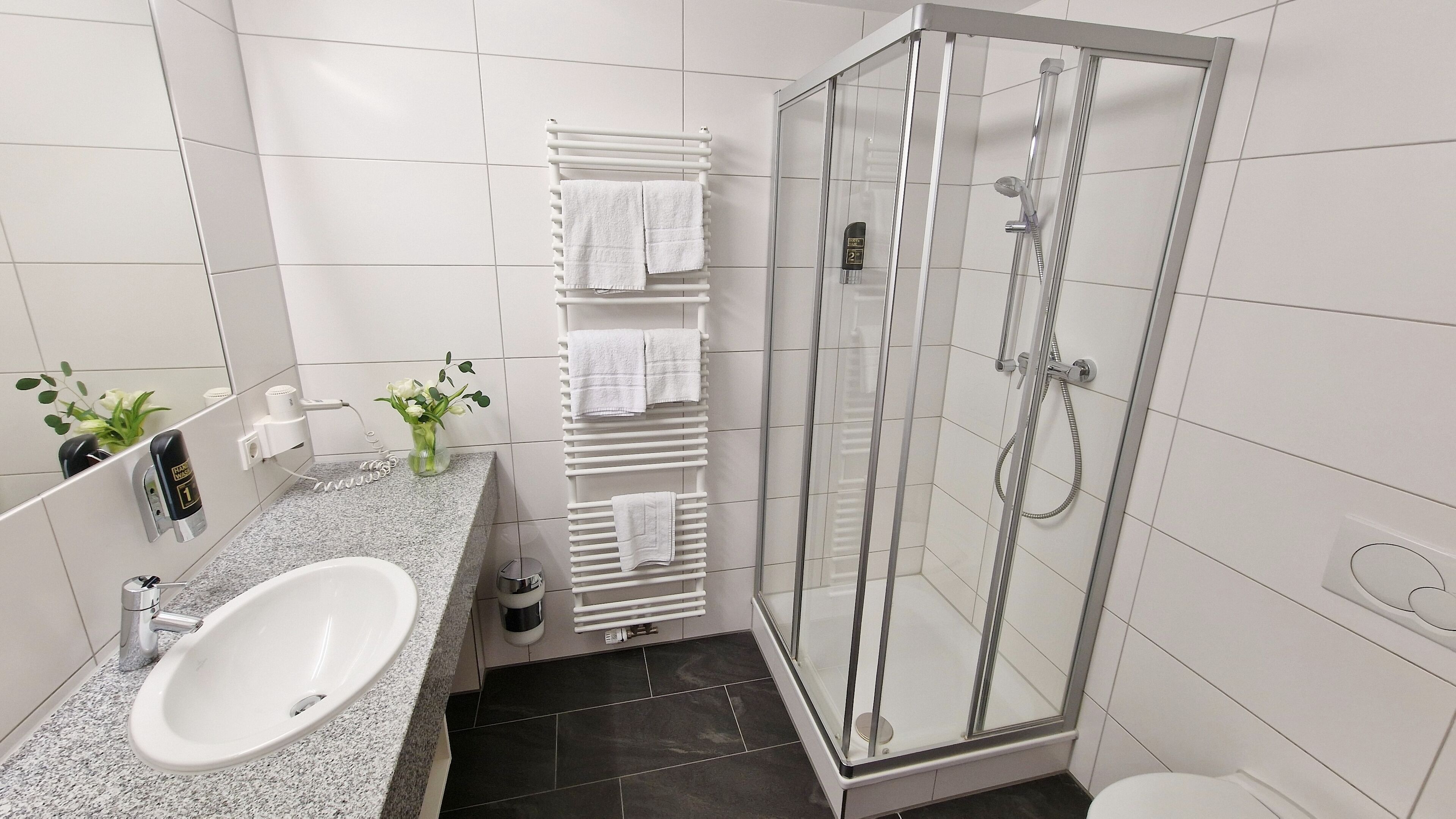 Ein modernes Badezimmer mit einer Duschkabine, einer Arbeitsplatte aus Granit, einem weißen Waschbecken und einem Handtuchhalter mit mehreren Handtüchern.