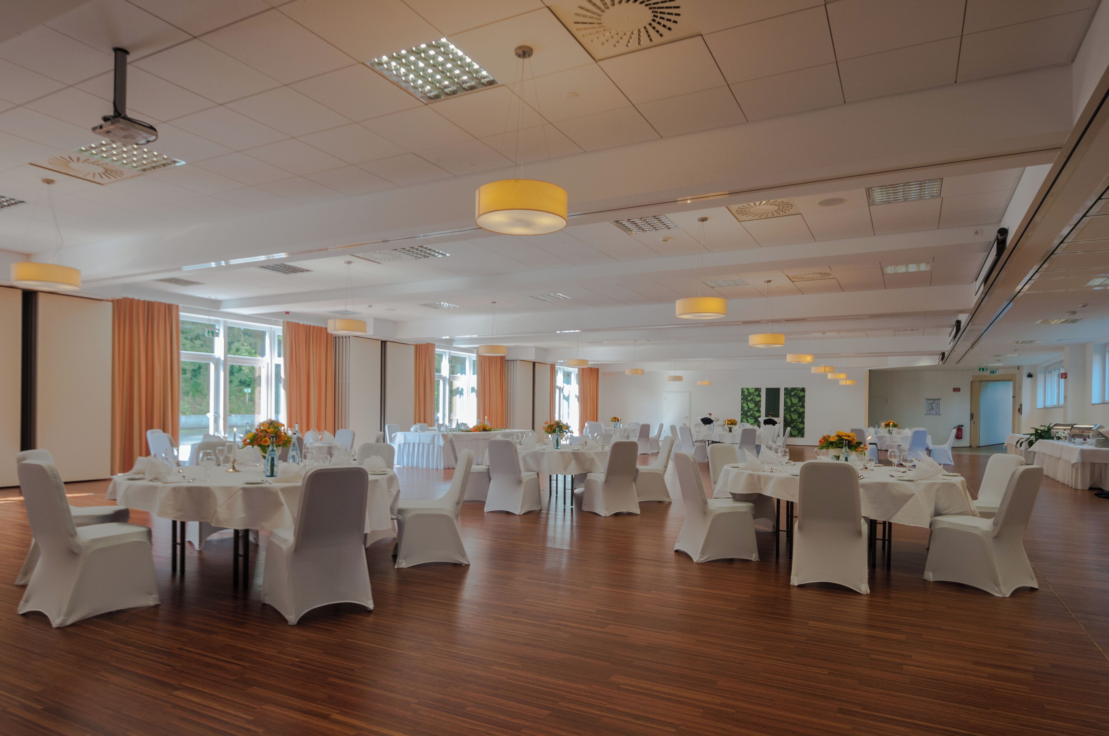 Un amplio salón de banquetes con varias mesas redondas, sillas blancas y lámparas colgantes amarillas.