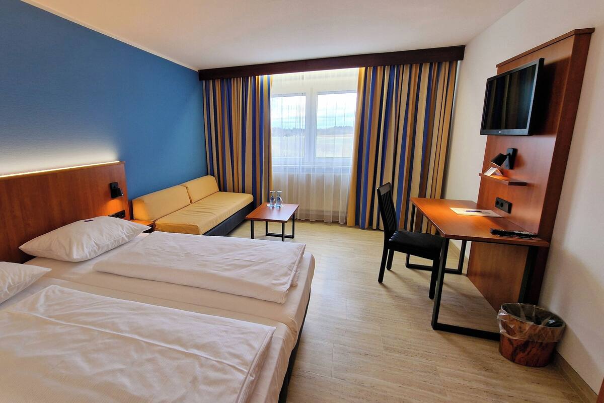 A hotel room with two beds, a sofa, a desk with a chair, a TV mounted on the wall, and a window with curtains.