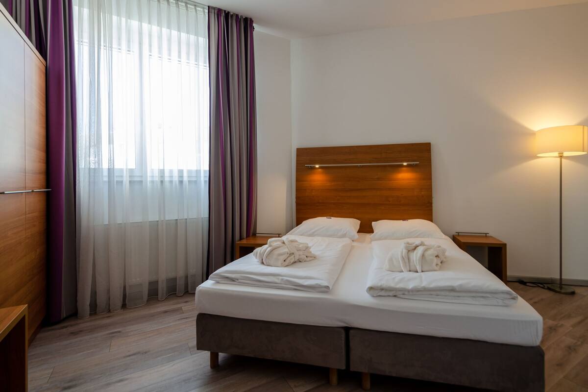 A neatly made double bed with a wooden headboard, two bedside tables, a floor lamp, and a large window with sheer curtains.