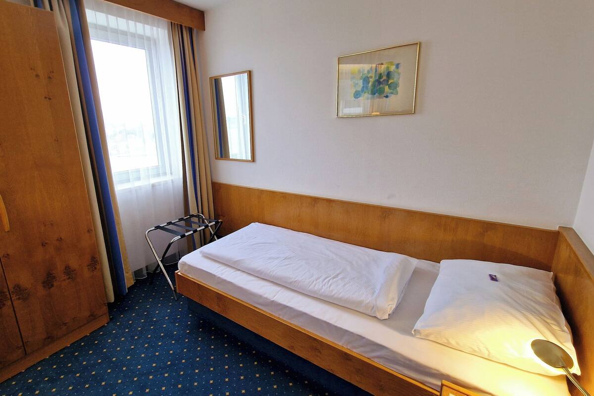 A hotel room with a single bed, a wardrobe, a window with curtains, and a framed picture on the wall.