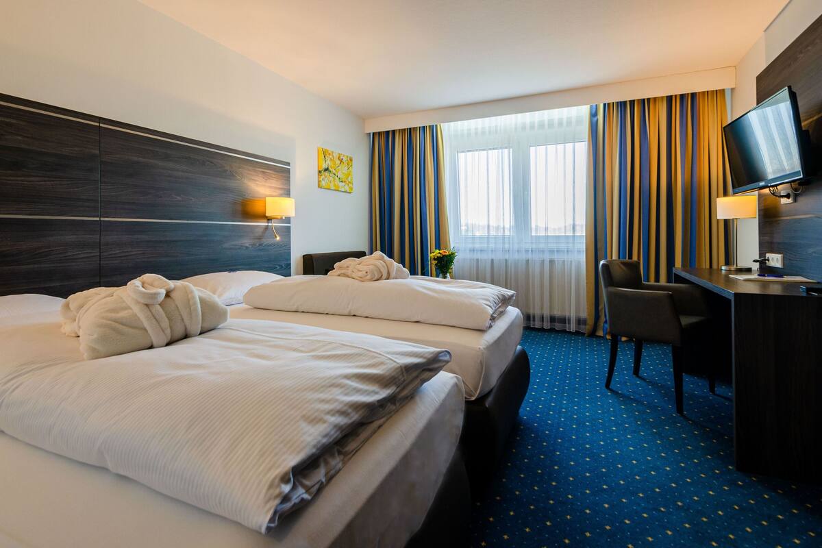 A hotel room with a large bed, a desk, a chair, a TV, and a window with curtains.