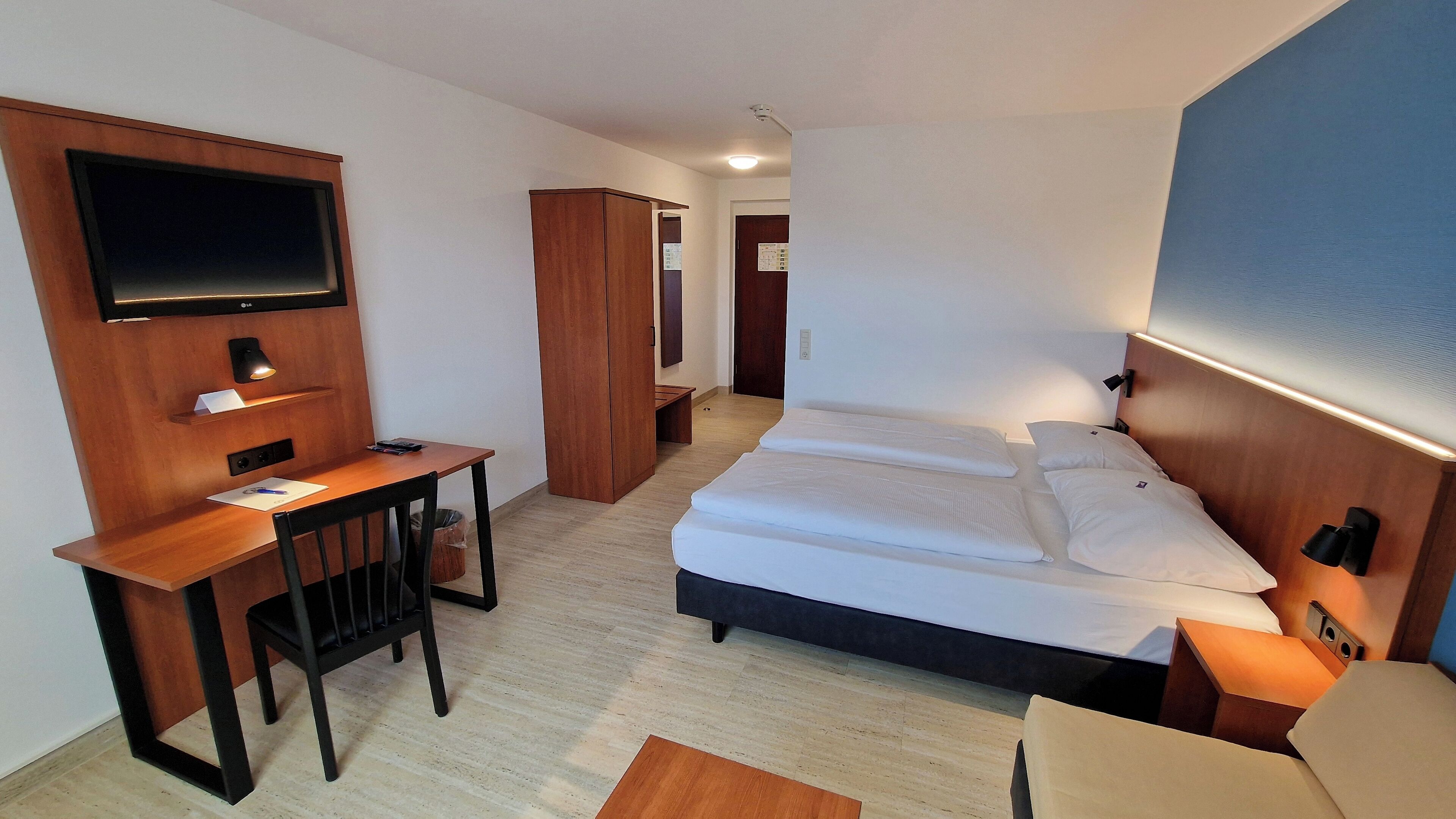 A hotel room with a bed, a desk, a chair, a TV mounted on the wall, and a wardrobe.