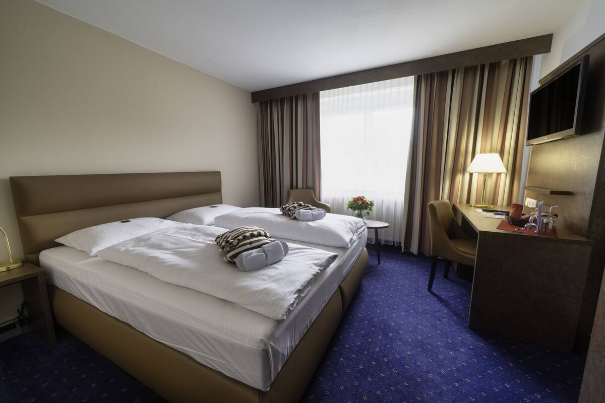 A hotel room with a large bed, a desk, a chair, a TV, and a window with curtains.