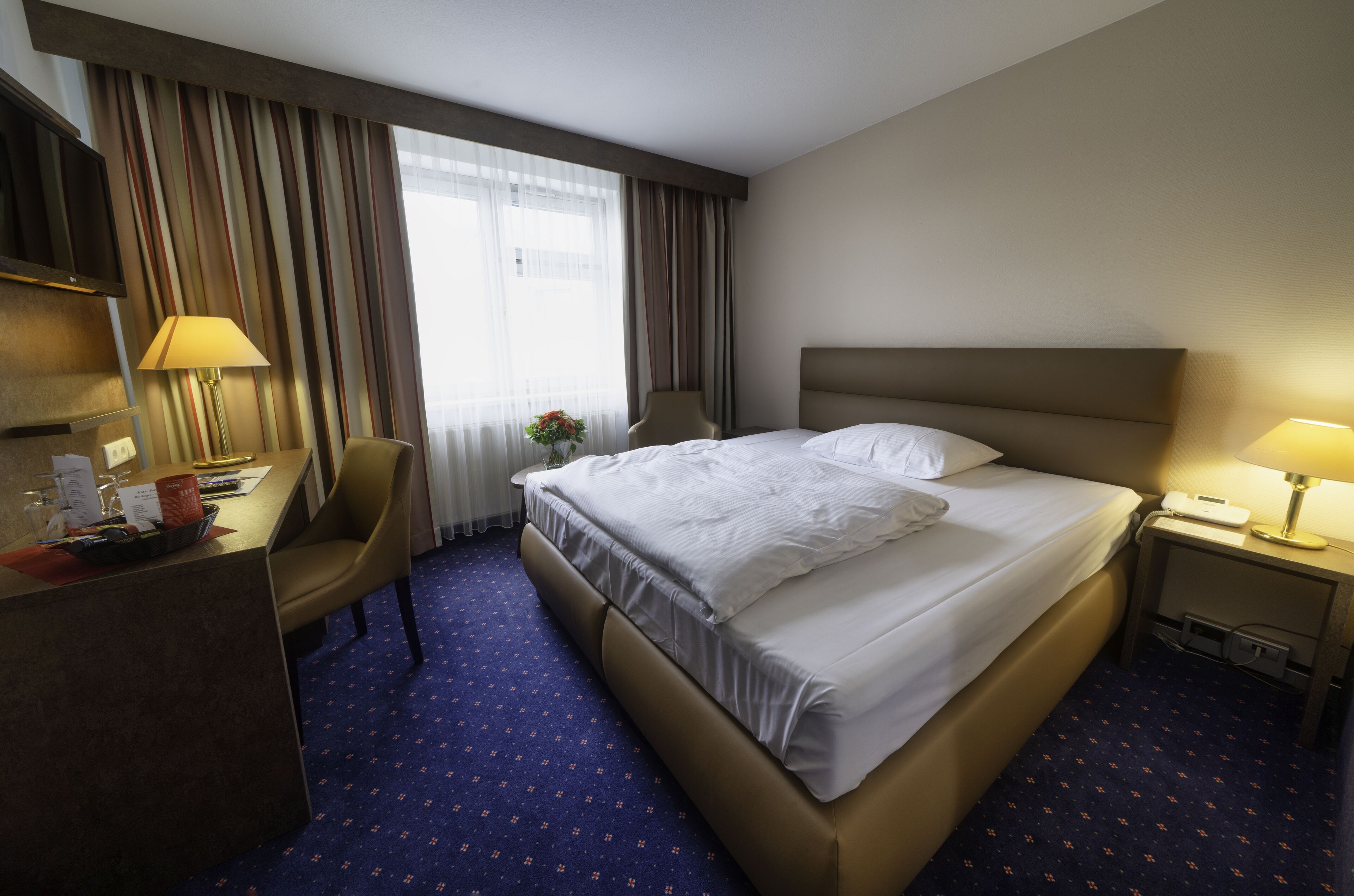Una habitación de hotel con una cama grande, un escritorio con lámpara, una silla y un televisor.