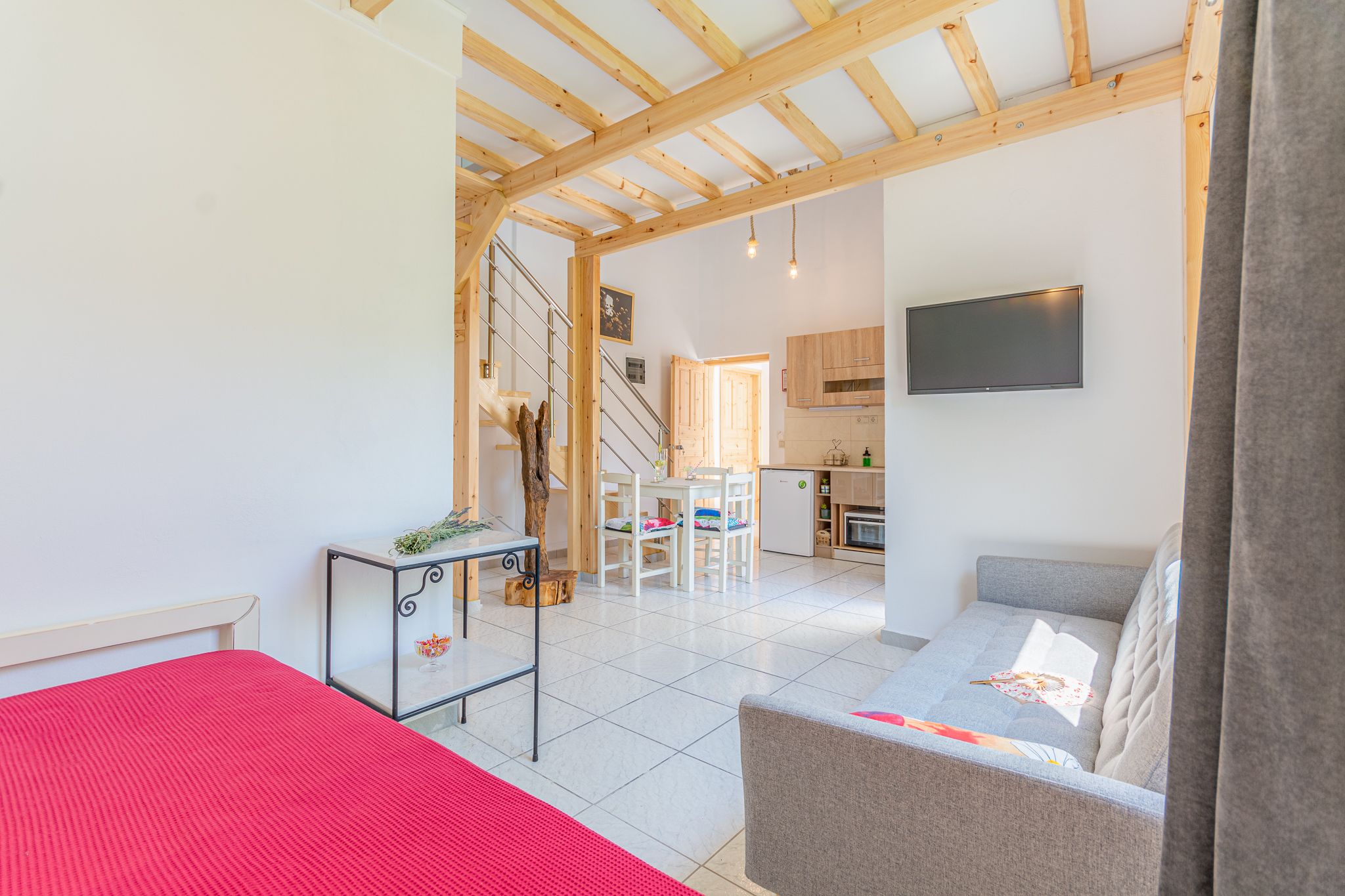 A compact living space with a bed, a sofa, a small table, a staircase leading to a kitchen, and a TV mounted on the wall.
